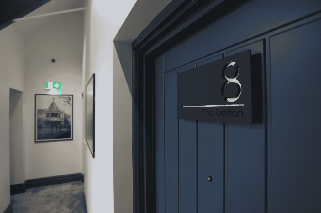 Apartment number 8 door, named 'The Dalton' and hallway, with framed pictures on the wall and an emergency exit sign.