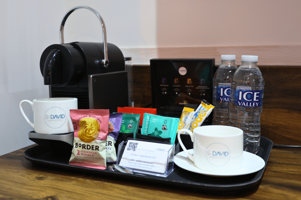 Drinks tray including a Nespresso coffee machine and box including pods and a description of them. Tea bags, biscuits, 2 bottles of water and 2 coffee cups are also on the tray. As well as business cards.