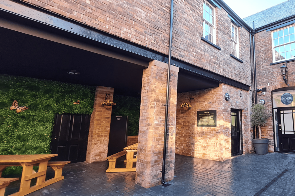 Stylish courtyard with a 'living wall' covered with miss and a vending machine in the corner. Wooden bench tables and hanging baskets complete the seating area and a plaque detailing the history of the building is attached to the wall.