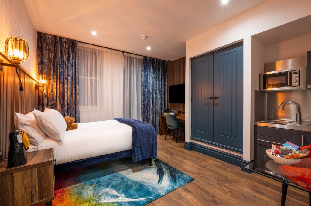 Modern hotel room with wooden slatted headboard, warm wall-mounted lights, double bed with white linens and navy throws, colorful rug, and a wooden desk with an orange chair, TV on the wall and kitchenette with sink, microwave and cupboards.