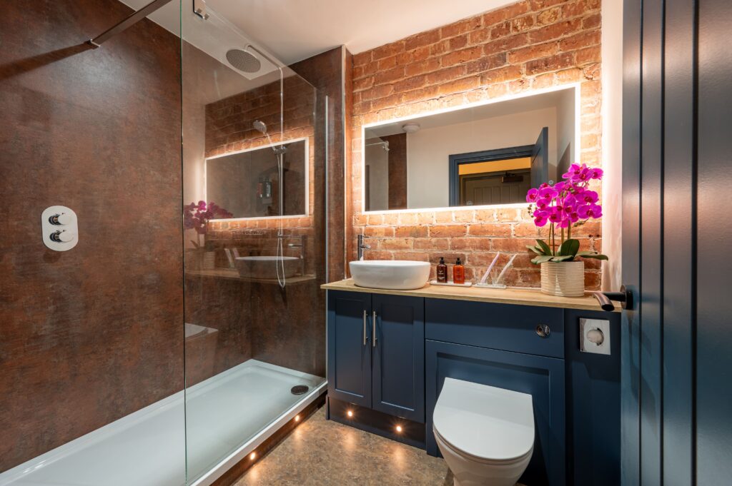 Modern bathroom with backlit mirror and exposed brick wall. Large, spacious shower cubicle and sleek cupboards, finished with floor lights and a toilet.