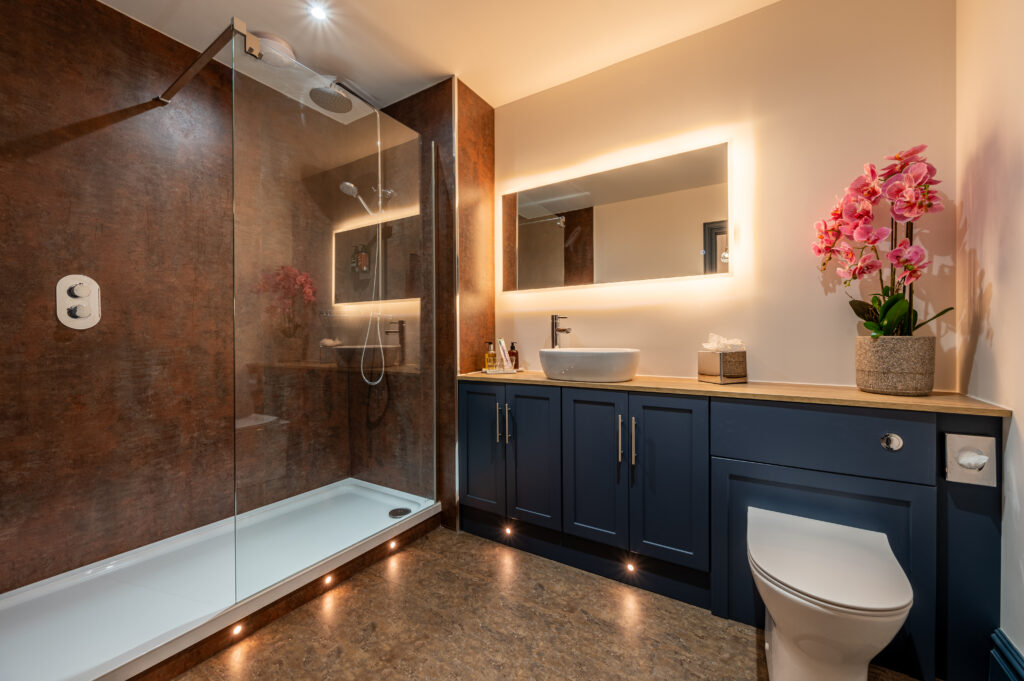 Modern bathroom with backlit mirror, large, spacious shower cubicle and sleek cupboards, finished with floor lights and a toilet.