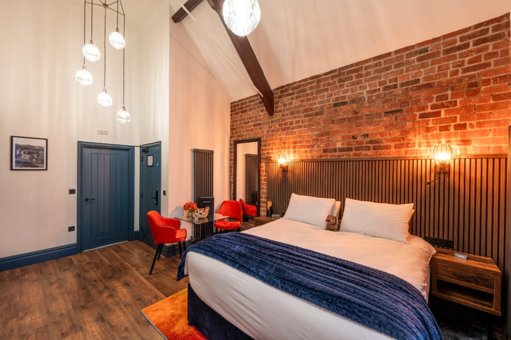 Modern hotel room with wooden slatted headboard, warm wall-mounted lights, double bed with white linens and navy throws, colorful rug, and a wooden desk with a orange chairs and an exposed brick wall at the back.