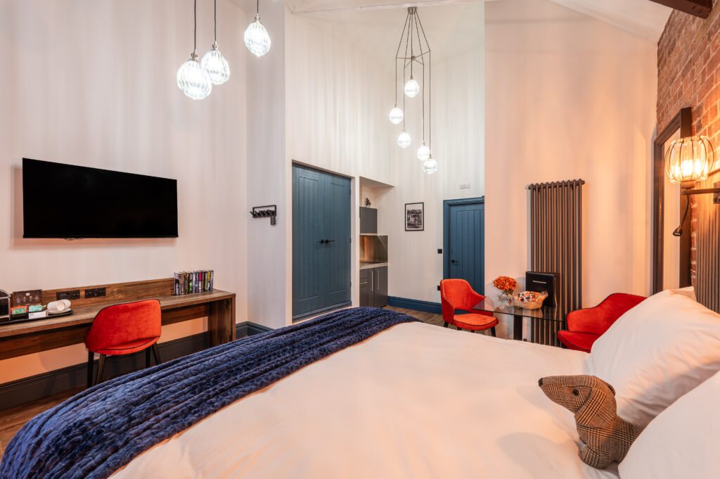 Modern twin hotel room with wooden slatted headboard, warm wall-mounted lights, double bed with white linens and navy throws, colorful rug, and a wooden desk with an orange chair and TV on the wall.