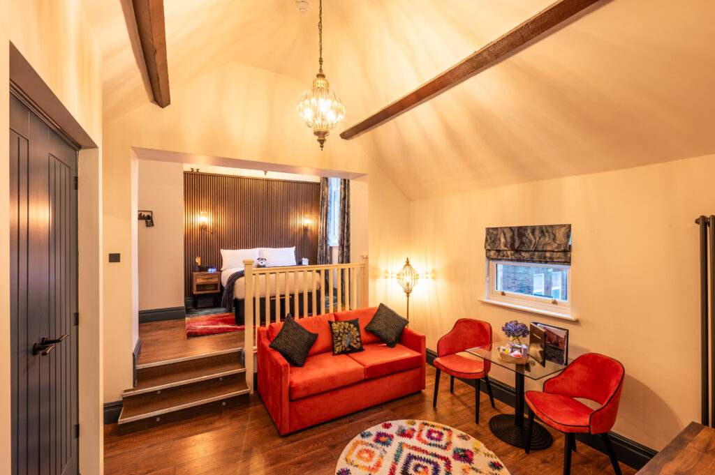 Cosy room with sofa, table and chairs with a double bed up some small stairs against a panelled wall. Large light fittings hang from the ceiling and emit a warm light.