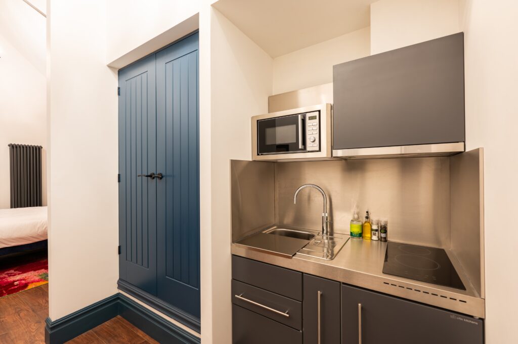 Compact kitchenette with stainless steel countertop, sink, electric stove, and microwave, adjacent to a blue double-door storage cabinet and white walls with dark trim.