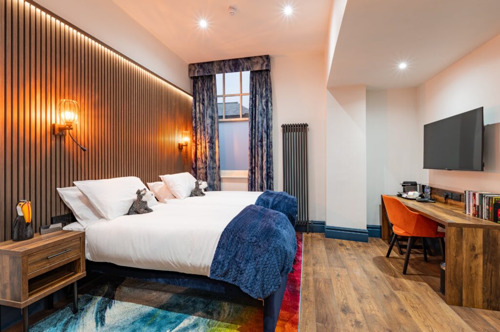 Modern twin hotel room with wooden slatted headboard, warm wall-mounted lights, two beds with white linens and navy throws, colorful rug, and a wooden desk with an orange chair and TV on the wall.