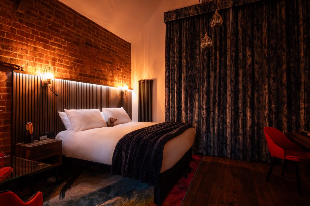 Cozy hotel room with exposed brick wall, warm ambient lighting, a large bed with white linens and a dark blanket, and elegant dark curtains, featuring modern decor and red accent chairs.